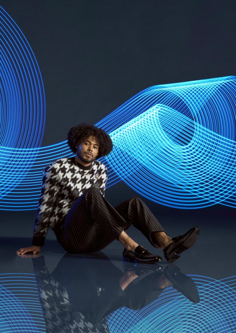 The featured image is a model wearing black Stacy Adams slip on shoes, black striped pants, and a white and black sweater while sitting on the floor with a blue abstract background.